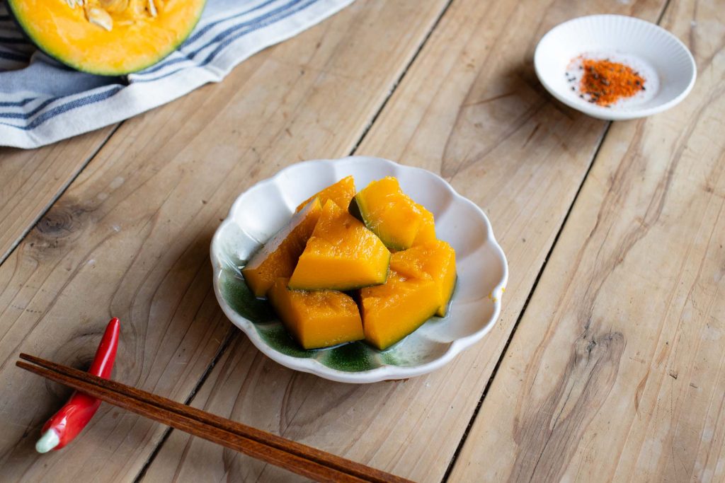 A small plate of simmered Japanese Pumpkin, chopsticks sitting on a chilli chopstick rest, a small plate of Japanese chilli spice mix and two quarters of uncooked Japanese Pumpkin on a striped dishcloth.