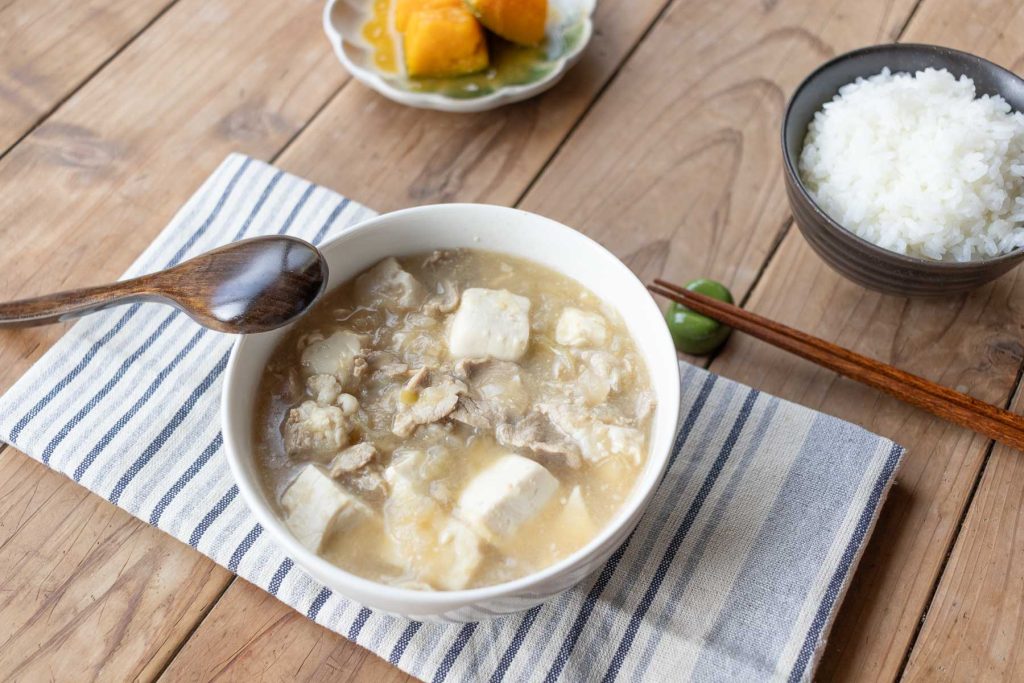A bowl of Pork Miso Soup (Tonjiru) on a tea towel with a bowl of rice and steamed kabocha on the side.