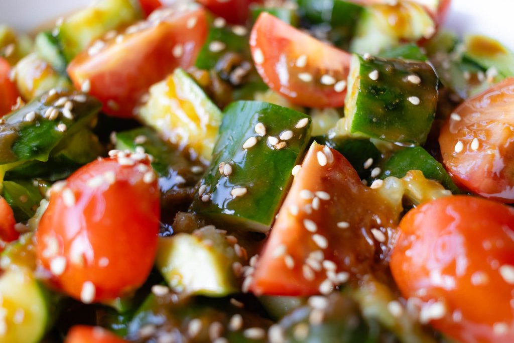 a close up of a plate of smashed sesame cucumber salad. There are cucumbers and tomatoes coated in a silky sauce and garnished with some sesame seeds.