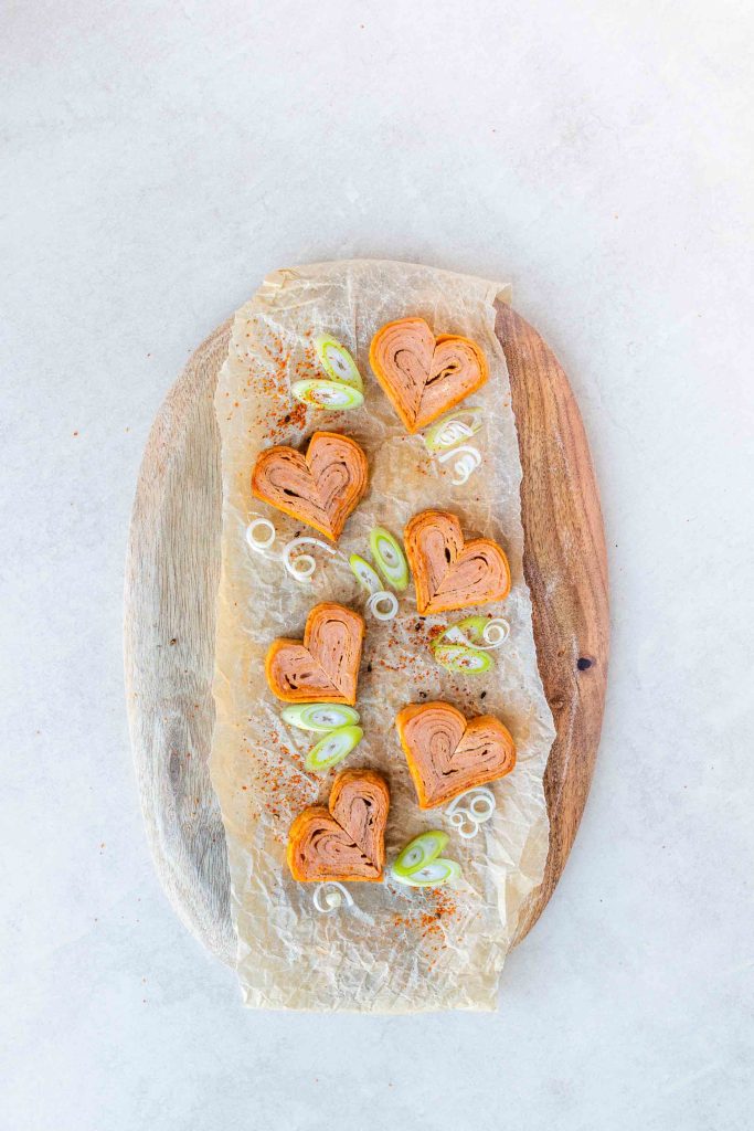 6 heart shaped gochujang Tamagoyaki pieces, garnished with spring onions, on a wooden chopping board.
