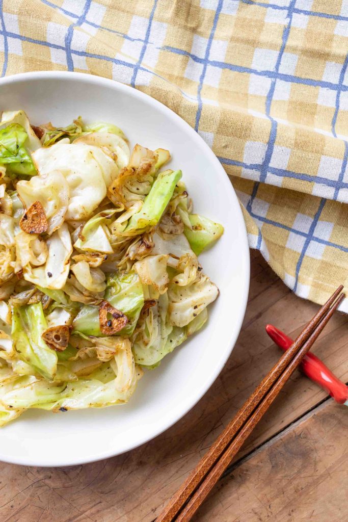 A plate of stir fried cabbage with pieces of garlic sitting on a yellow and blue checkerboard cloth and to the side a pair of chopsticks on a chilli chopstick holder.