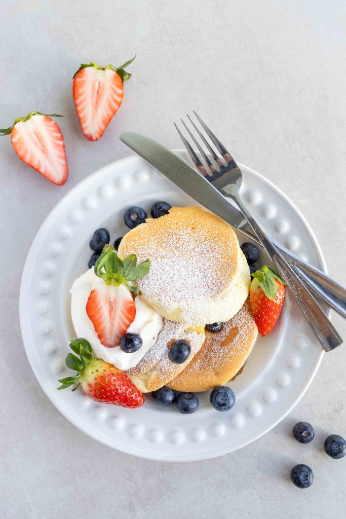 A plate of Japanese Soufflé Pancakes, which requires no mixer, with icing sugar sprinkled on top, whipped cream, strawberries and blueberries.