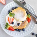 A plate of Japanese Soufflé Pancakes, which requires no mixer, with icing sugar sprinkled on top, whipped cream, strawberries and blueberries.