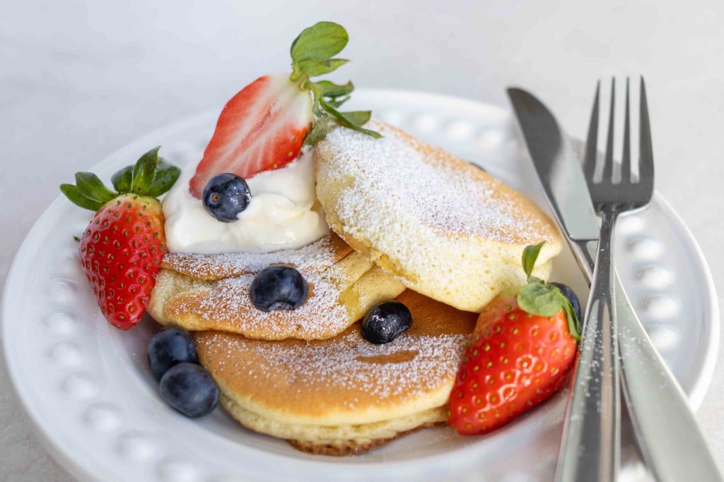 A plate of Japanese Soufflé Pancakes, which requires no mixer, with icing sugar sprinkled on top, whipped cream, strawberries and blueberries.