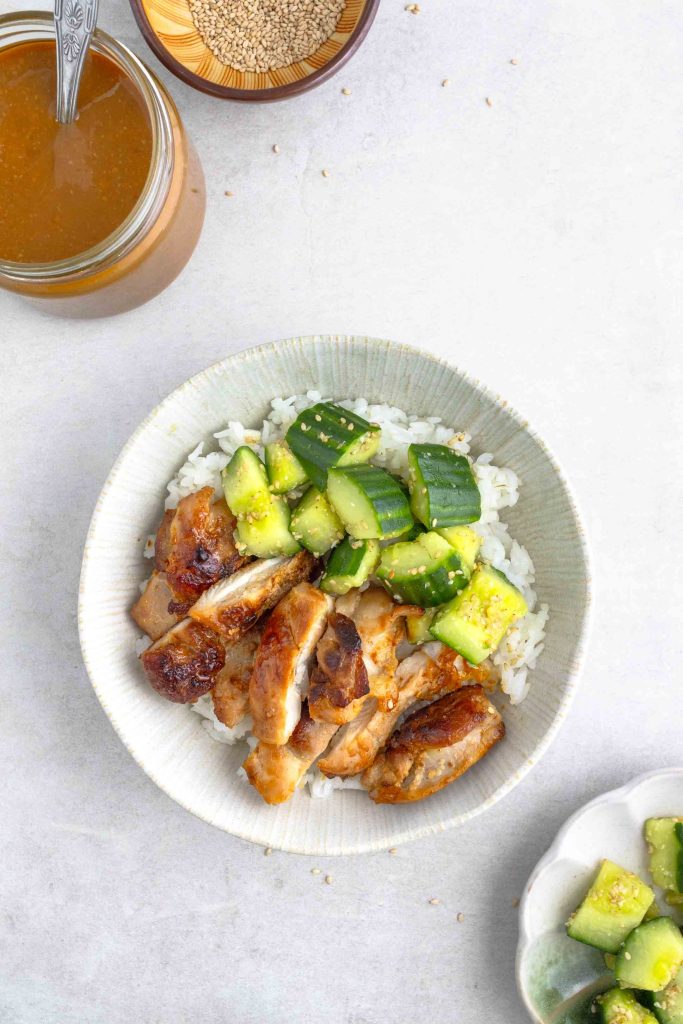 A bowl of miso chicken thighs chopped up with some smashed cucumbers, all on top of some white rice. There is also a pot of miso marinade in the top left corner.