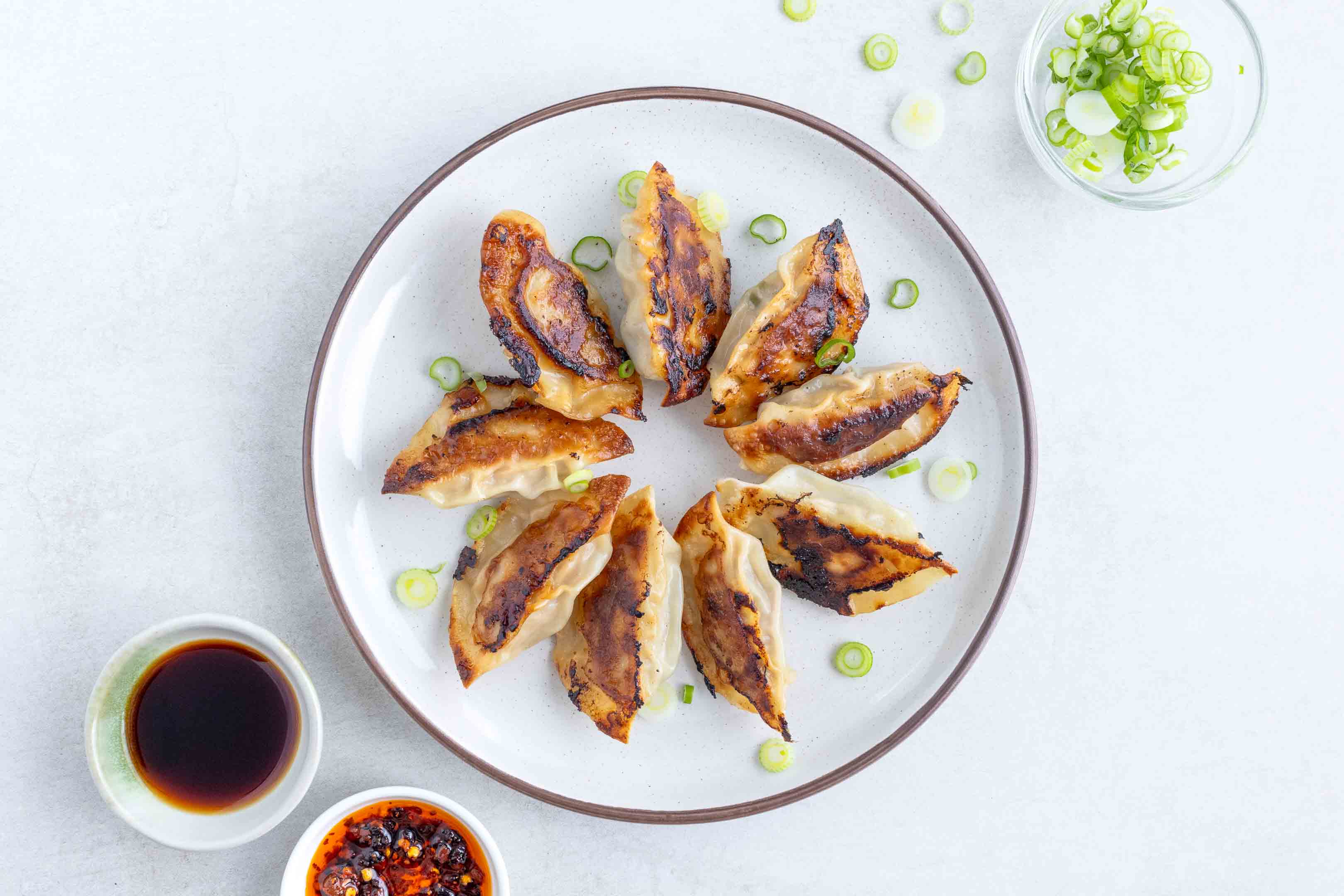 A plate of Gyoza with a side of dipping sauce and chilli oil. Sprinkled with some spring onion.